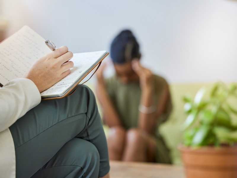 Therapy writing, psychologist and sad black woman with stress, headache and burnout in office. Support, therapist consultation and mental health check with psychology work of a patient on a sofa.